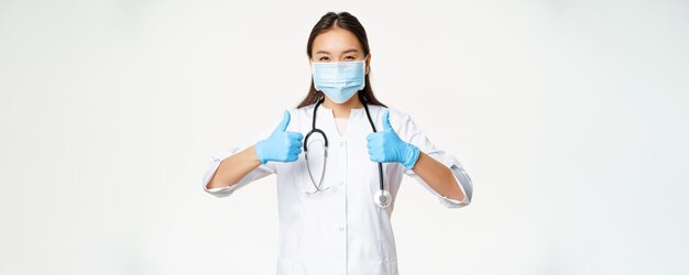 Free photo healthcare and covid19 preventive measures concept asian physician in face mask and rubber gloves shows thumbs up stands in uniform over white background