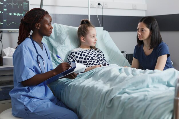 病室での回復の成功についてメモを取っている医療小児科クリニックの看護師。回復病棟にいる間、母親は少女の健康被害について話す看護師に注意を払っています。