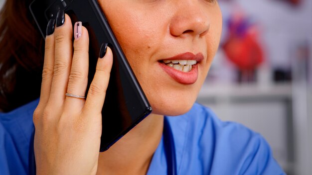 医療ユニフォームを着た病院で電話を使用して遠隔地の患者に相談する医療医師。遠隔医療コミュニケーション、診断で患者を支援する医師助手のクローズアップ