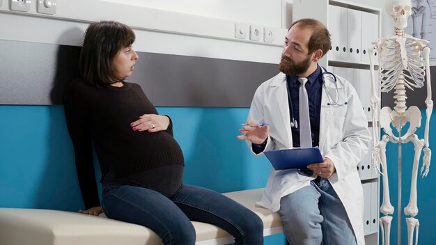 건강 관리 전문가는 의료 캐비닛에서 임신한 여성을 상담하고 검진 방문 시 메모를 합니다. 남성 의사는 임신 배가 있는 환자와 이야기를 나누며 출산을 준비합니다.