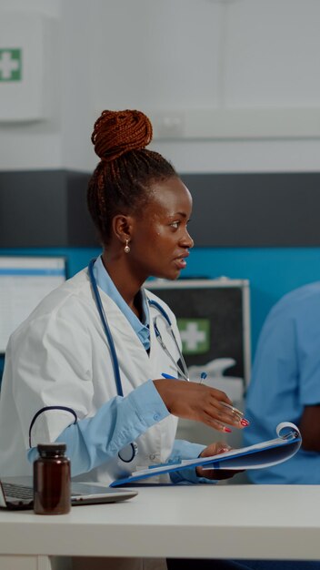 Healthcare specialist discussing with old patient at desk in medical office. Doctor checking disease symptoms for senior person while nurse and elder woman sitting in background