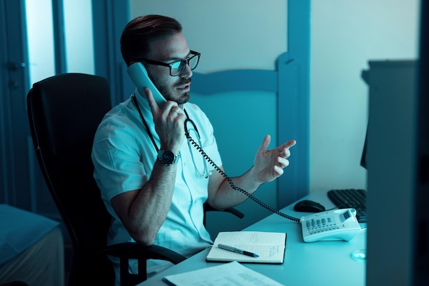 Free photo healthcare worker sitting in medical examination room and talking over the phone with someone