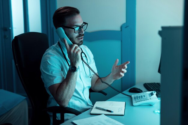 Healthcare worker sitting in medical examination room and talking over the phone with someone