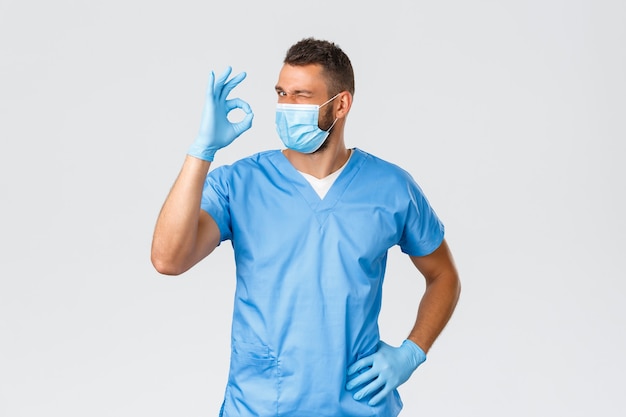 医療従事者、covid-19、コロナウイルスおよび予防ウイルスの概念。スクラブと医療マスクの生意気なハンサムな医者は、最高のサービス品質を保証し、保証し、大丈夫なサインとウィンクを示します