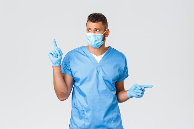 Healthcare workers, covid-19, coronavirus and preventing virus concept. Concerned and distressed handsome doctor, nurse or intern in scrubs and medical mask, look left, pointing sideways