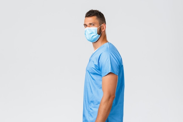 Healthcare workers, covid-19, coronavirus and preventing virus concept. Confident handsome doctor, male nurse in scrubs and medical mask standing profile, turning camera with determined look