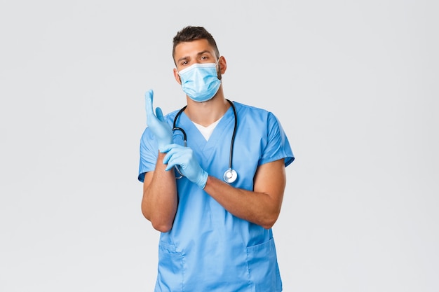 Healthcare workers, covid-19, coronavirus and preventing virus concept. Confident serious-looking male doctor, nurse in medical mask and scrubs, put on gloves to observe patient in clinic.