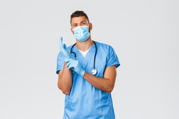 Healthcare workers, covid-19, coronavirus and preventing virus concept. Confident serious-looking male doctor, nurse in medical mask and scrubs, put on gloves to observe patient in clinic.