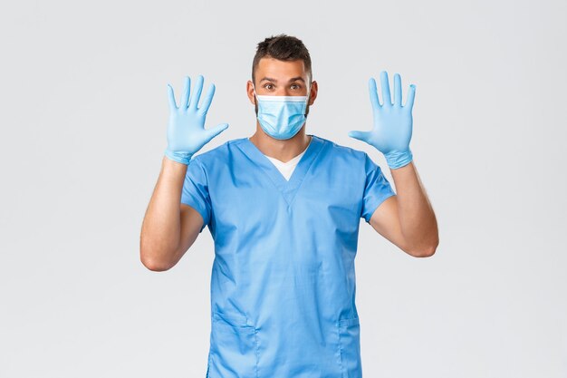Healthcare workers, covid-19, coronavirus and preventing virus concept. Excited smiling male doctor, nurse in medical mask and gloves, raising hands up wear rubber gloves, grey background