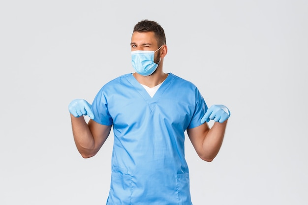 Healthcare workers, covid-19, coronavirus and preventing virus concept. Handsome cheeky doctor invite take test screening at their lab clinic, nurse in scrubs wink camera pointing down