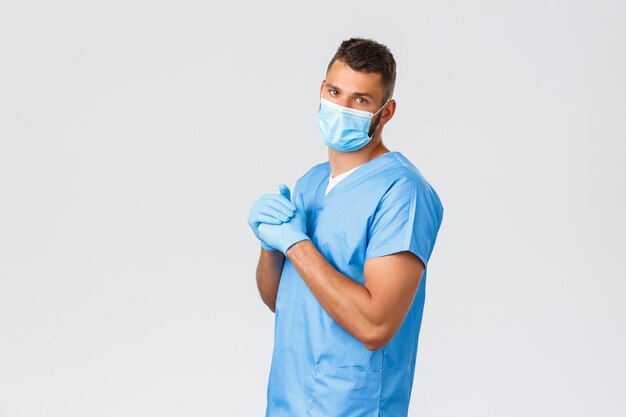 Healthcare workers, covid-19, coronavirus and preventing virus concept. Handsome doctor, male nurse in medical mask and scrubs, crack knuckles, night shift at clinic, grey background