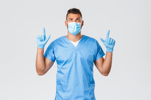 의료 종사자, covid-19, 코로나바이러스 및 예방 바이러스 개념. 잘 생긴 진지한 남성 의사, 수술용 마스크와 의료용 마스크를 쓴 간호사, 찾고 손가락을 위로 가리키며 배너 프로모션을 읽고 있습니다.