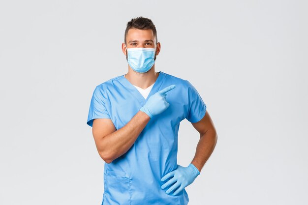 Healthcare workers, covid-19, coronavirus and preventing virus concept. Handsome smiling doctor, male nurse in medical mask and scrubs, pointing finger right, showing clinic banner, promo.