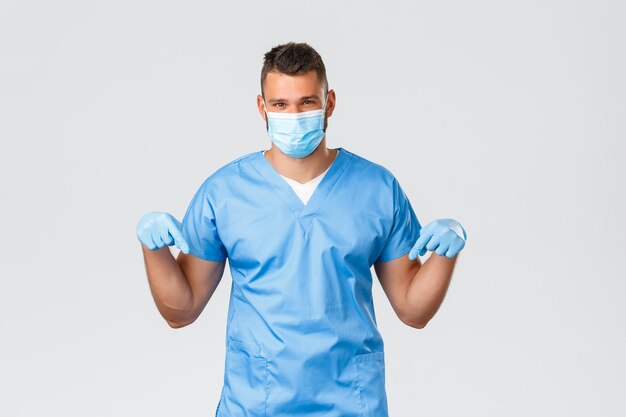 의료 종사자, covid-19, 코로나바이러스 및 예방 바이러스 개념. 의료용 마스크와 스크럽을 쓴 잘생긴 남자 의사는 손가락을 아래로 가리키며 특별 프로모션, 좋은 소식을 알려줍니다.