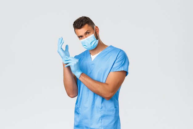 Healthcare workers, covid-19, coronavirus and preventing virus concept. Handsome young professional doctor, male nurse in medical mask and scrubs, put on rubber gloves to observe patient