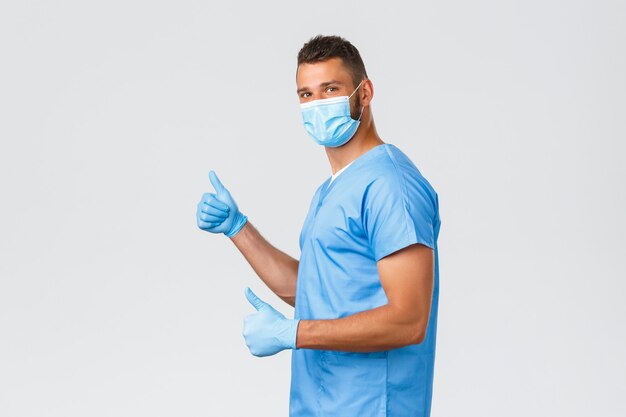 Медицинские работники, covid-19, коронавирус и концепция профилактики вирусов. Оптимистичный жизнерадостный мужчина-врач, медсестра или интерн в скрабах, перчатках и медицинской маске, показывает вверх большие пальцы в поддержку или одобрение