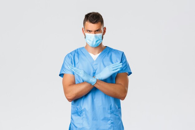 Медицинские работники, covid-19, коронавирус и концепция профилактики вирусов. Серьезно злой и уверенный в себе медсестра или врач, носить медицинскую маску и перчатки, делать крестик, запрещающий или запрещающий.