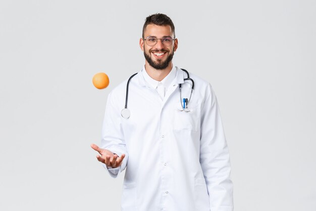 Foto gratuita operatori sanitari, assicurazione medica, laboratorio clinico e concetto di covid-19. medico ispanico sorridente allegro, medico in camice bianco, lancia l'arancia, consiglia di mangiare frutta sana con vitamine