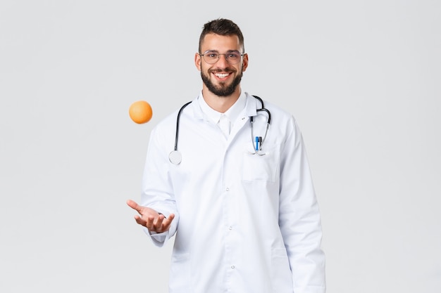 医療従事者、医療保険、クリニックラボ、covid-19コンセプト。陽気な笑顔のヒスパニック系医師、白衣を着た医師、オレンジを投げ、健康的なビタミンフルーツを食べることをお勧めします