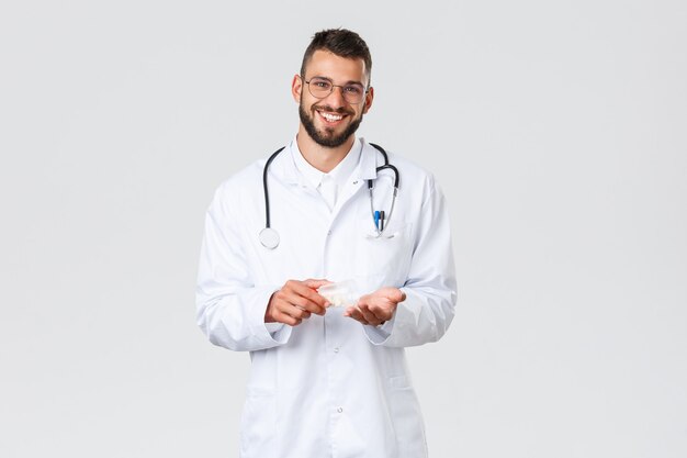 Медицинские работники, медицинское страхование, концепция пандемии и covid-19. Красивый улыбающийся врач в белом халате и очках, держа в руке лекарство, советует использовать лекарства или антибиотики.