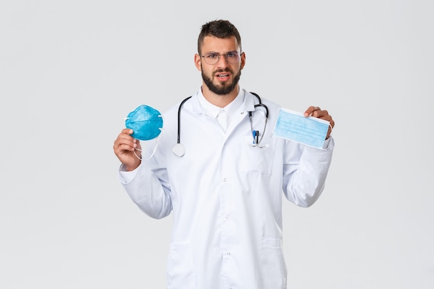 Медицинские работники, медицинское страхование, концепция пандемии и covid-19. Озадаченный молодой красивый доктор в белом халате, очки, показывающие респиратор и медицинскую маску, два разных СИЗ.