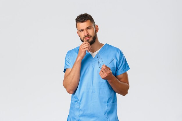 Healthcare workers, medicine, covid-19 and pandemic self-quarantine concept. Serious and thoughtful smart male nurse, doctor in blue scrubs, rub beard and look interested, grey background