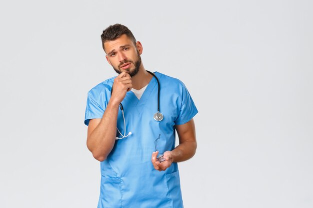 의료 종사자, 의학, covid-19 및 전염병 자가 격리 개념. 파란색 수술복을 입은 피곤한 젊은 의사, 남성 간호사가 걱정하는 환자의 말을 듣고, 피로가 거의 없는 집중된 얼굴