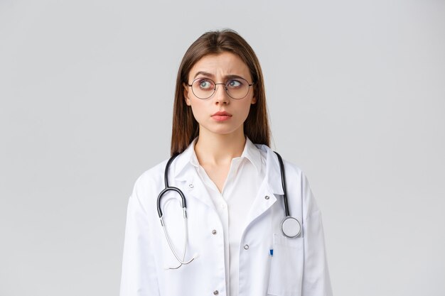 의료 종사자, 의학, 보험 및 covid-19 전염병 개념. 의사는 이상한 것을 봅니다. 흰색 스크럽, 안경, 인상을 찌푸리고 왼쪽을 응시하는 혼란스럽고 수상한 여성 의사