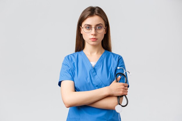 의료 종사자, 의학, 보험 및 covid-19 전염병 개념. 진지하고 단호한 전문 여성 간호사, 파란색 수술복을 입은 의사, 청진기를 들고 안경을 쓰고 자신감을 보입니다.