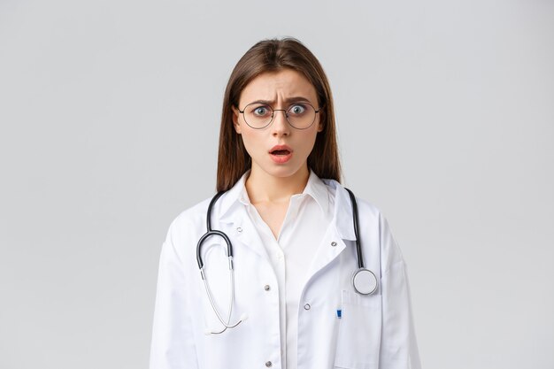 의료 종사자, 의학, 보험 및 covid-19 전염병 개념. 흰색 수술복과 안경을 쓴 신경질적인 여성 의사, 의사가 헐떡거리며 걱정하는 카메라를 쳐다보고 있다