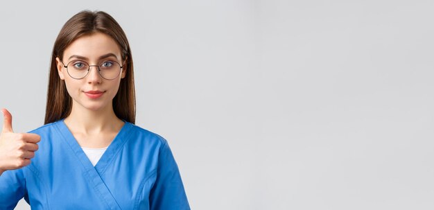 Healthcare workers medicine insurance and covid19 pandemic concept Closeup supportive and confident female nurse doctor in blue scrubs and glasses show approval thumbsup guarantee