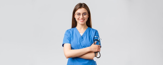 医療従事者の医療保険とcovid19パンデミックの概念青いスクラブとメガネで自信を持って熱心な若い女性看護師の医者は聴診器の笑顔のカメラを喜ばせます