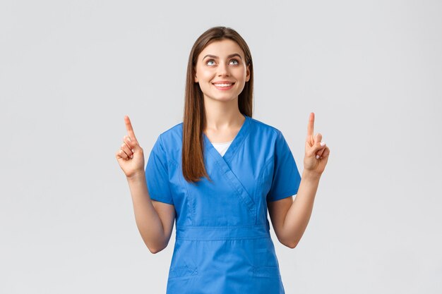 Healthcare workers, prevent virus, covid-19 test screening, medicine concept. Attractive female nurse or doctor in blue scrubs, pointing fingers and looking up with pleased smile, reading banner