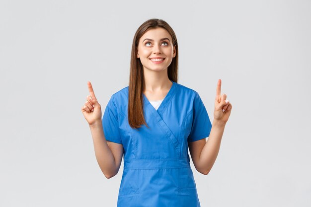 医療従事者、ウイルスの予防、covid-19検査スクリーニング、薬の概念。青いスクラブで魅力的な女性看護師または医師、指を指して、満足のいく笑顔で見上げる、バナーを読む