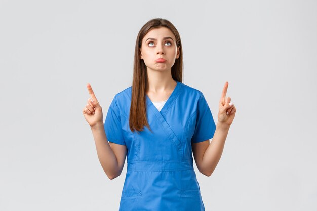 医療従事者、ウイルスの予防、covid-19検査スクリーニング、薬の概念。愚かな動揺かわいい医者、青いスクラブの女性看護師はがっかりし、指さし、見上げると不機嫌になります