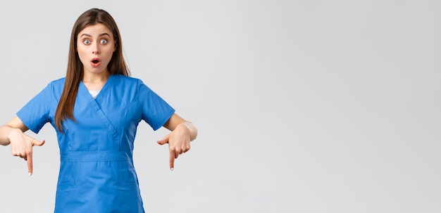 Healthcare workers prevent virus covid19 test screening medicine concept Shocked and surprised female nurse or doctor in blue scrubs pointing fingers down to inform people about promo