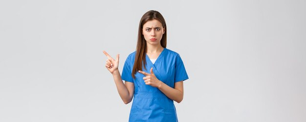 Healthcare workers prevent virus insurance and medicine concept Concerned and alarmed young female doctor or nurse in blue scrubs frowning anxious pointing fingers left at important info