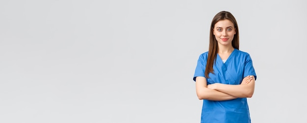 의료 종사자들은 바이러스 보험 및 의학 개념을 예방합니다. 파란색 수술복을 입은 자신감 넘치는 웃는 여성 간호사 의사가 팔짱을 끼고 단호한 표정으로 covid19 발병과 싸우고 있습니다.