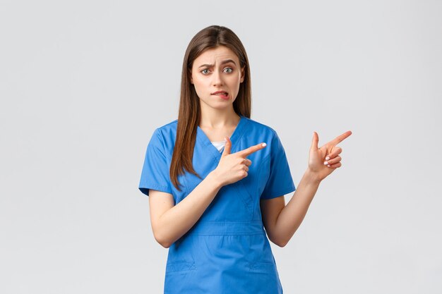 Healthcare workers, prevent virus, insurance and medicine concept. Nervous female intern worried standing in hospital. Doctor or nurse nervously biting lip and pointing fingers right