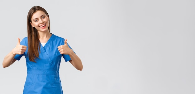 Healthcare workers prevent virus insurance and medicine concept Optimistic and enthusiastic female nurse doctor in blue scrubs thumbsup and smiling encourage patients stay home