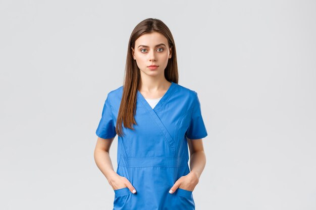 Healthcare workers prevent virus insurance and medicine concept Seriouslooking confident young medical worker nurse or doctor in blue scrubs hold hands in pockets and looking camera