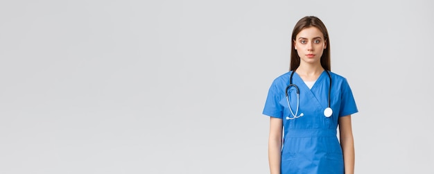 医療従事者はウイルス保険と薬の概念を防ぎます青いスクラブと聴診器で真面目な若い看護師の医者が灰色の背景に立っているカメラを見て