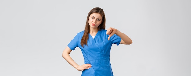 Healthcare workers prevent virus insurance and medicine concept Skeptical and disappointed young female nurse doctor in scrubs thumbdown and smirk rate bad idea judging awful behaviour