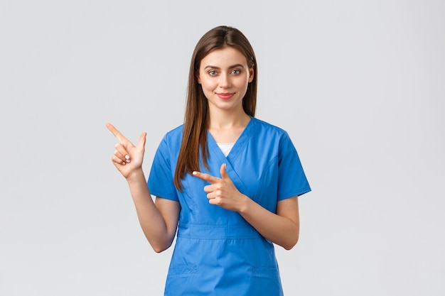医療従事者は、ウイルス、保険、薬の概念を防ぎます。患者の広告、重要な情報を表示するために左の指を指している青いスクラブで魅力的な女性医師または看護師の笑顔