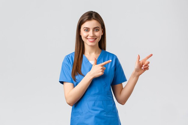 Healthcare workers, prevent virus, insurance and medicine concept. Smiling nurse or female doctor in blue scrubs, pointing fingers down, inform about coronavirus news, show information