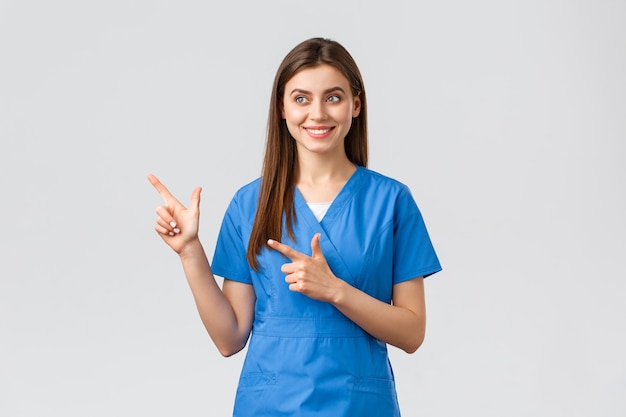 의료 종사자들은 바이러스, 보험, 의학 개념을 예방합니다. 파란 수술복을 입은 예쁜 간호사나 의사가 웃고 있고, 손가락을 왼쪽으로 가리키고 만족스러운 미소, 회색 배경으로 baner를 바라보고 있습니다.