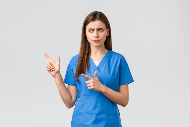 Healthcare workers, prevent virus, insurance and medicine concept. Something wrong. Serious displeased nurse or doctor in blue scrubs, frowning looking and pointing left concerned