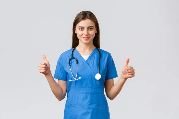 Healthcare workers, prevent virus, insurance and medicine concept. Supportive professional female nurse or doctor in blue scrubs, stethoscope, show thumbs-up in approval, smiling