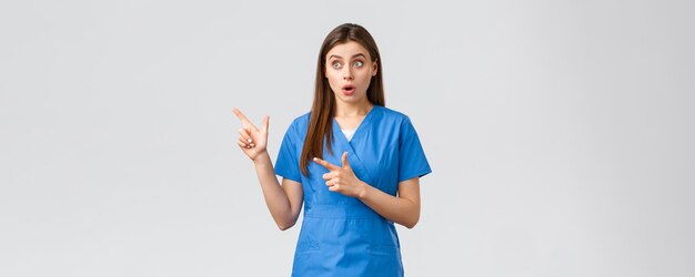 Healthcare workers prevent virus insurance and medicine concept Surprised female nurse or doctor in blue scrubs looking and pointing fingers upper left corner gasping amazed