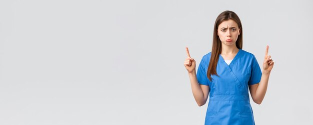 Healthcare workers prevent virus insurance and medicine concept Upset sobbing cute nurse or doctor in blue scrubs pouting feel uneasy pointing fingers up at unpleasant bad news show banner